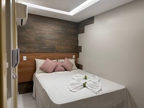 A modern hotel room with a wooden headboard, a neatly made bed with white linens, and a towel rack with folded towels. - Pousada Beija Flor (Maceió)