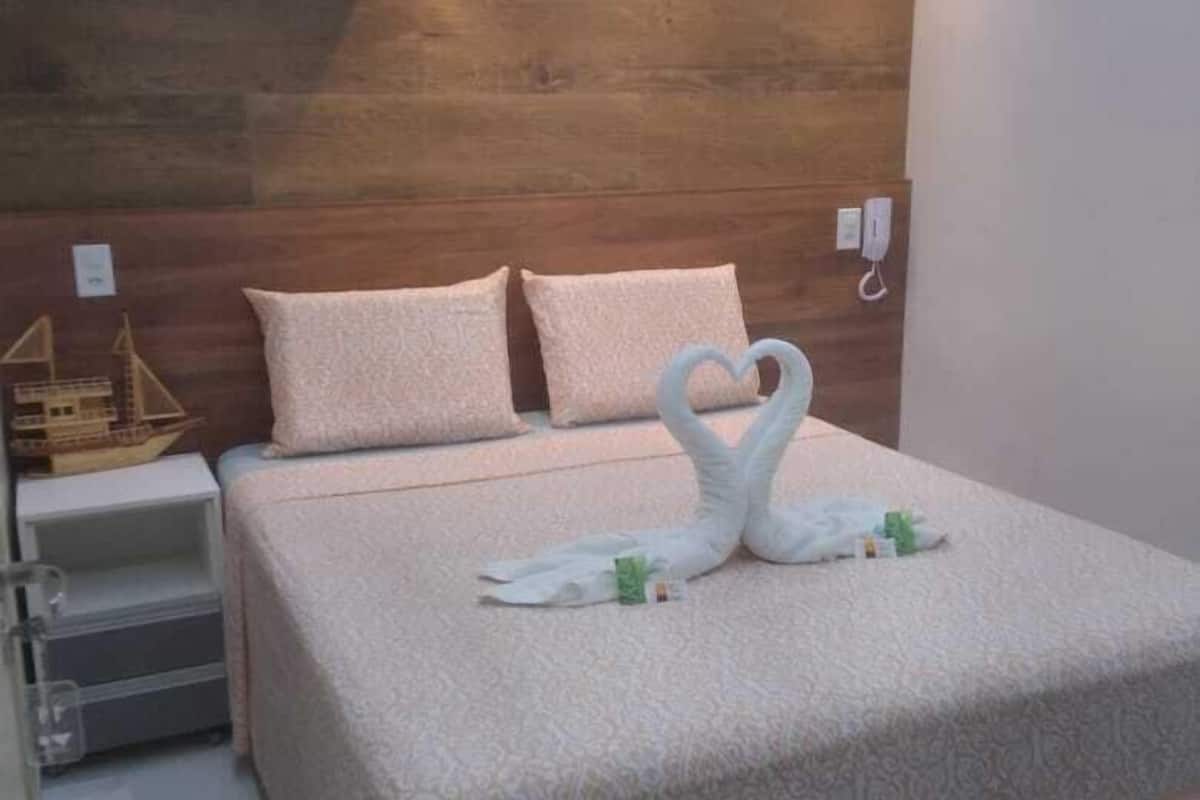 A neatly made bed with a heart-shaped towel arrangement, a wooden headboard, and a bedside table with a lamp.