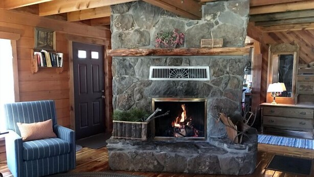 A stone fireplace with a fire, a striped armchair, and wooden walls. - Greer Pershing Cabin in the Meadow (Greer)