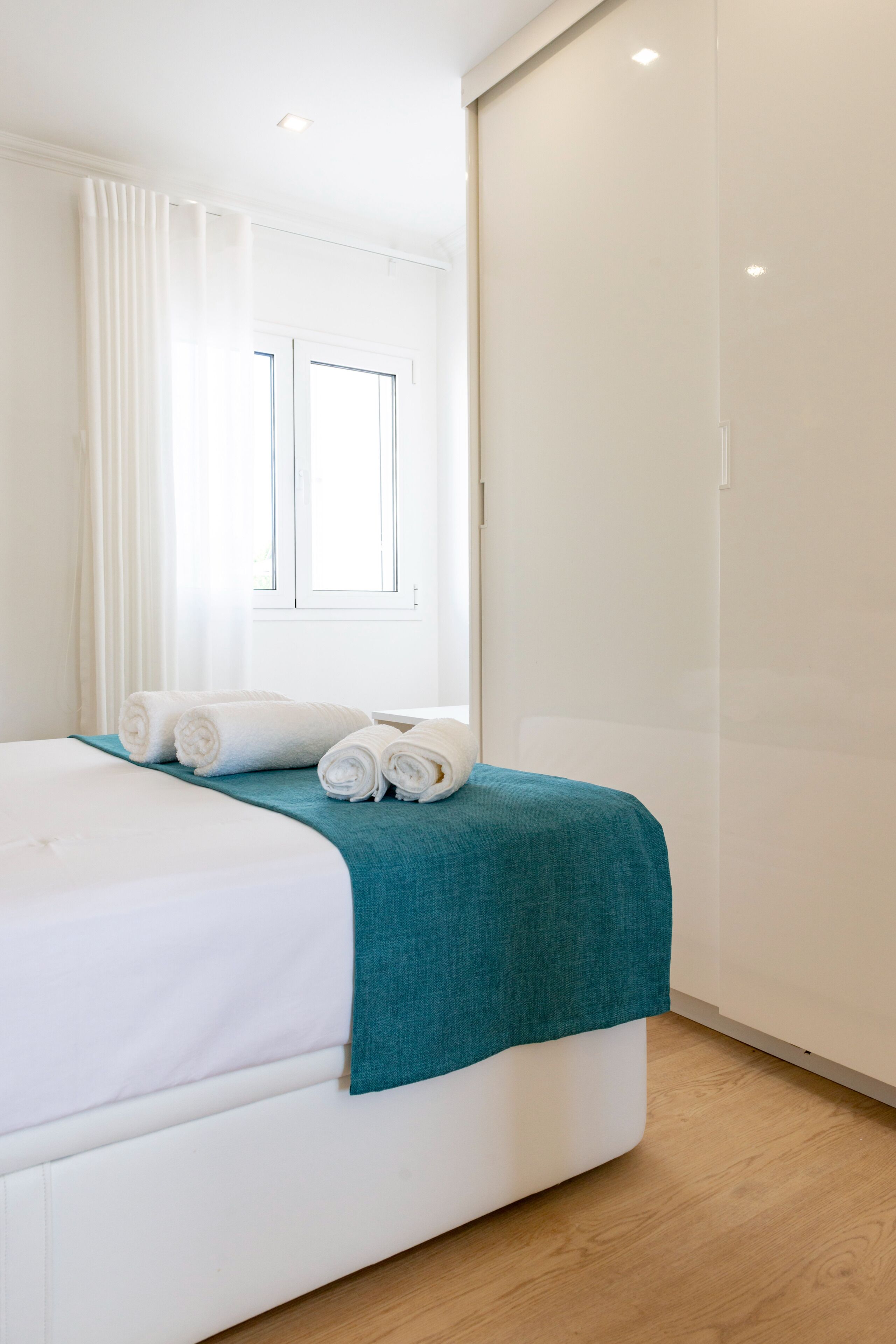 Un lit bien fait, avec du linge de lit blanc et une couverture turquoise, une fenêtre ornée de rideaux blancs et un plancher en bois.