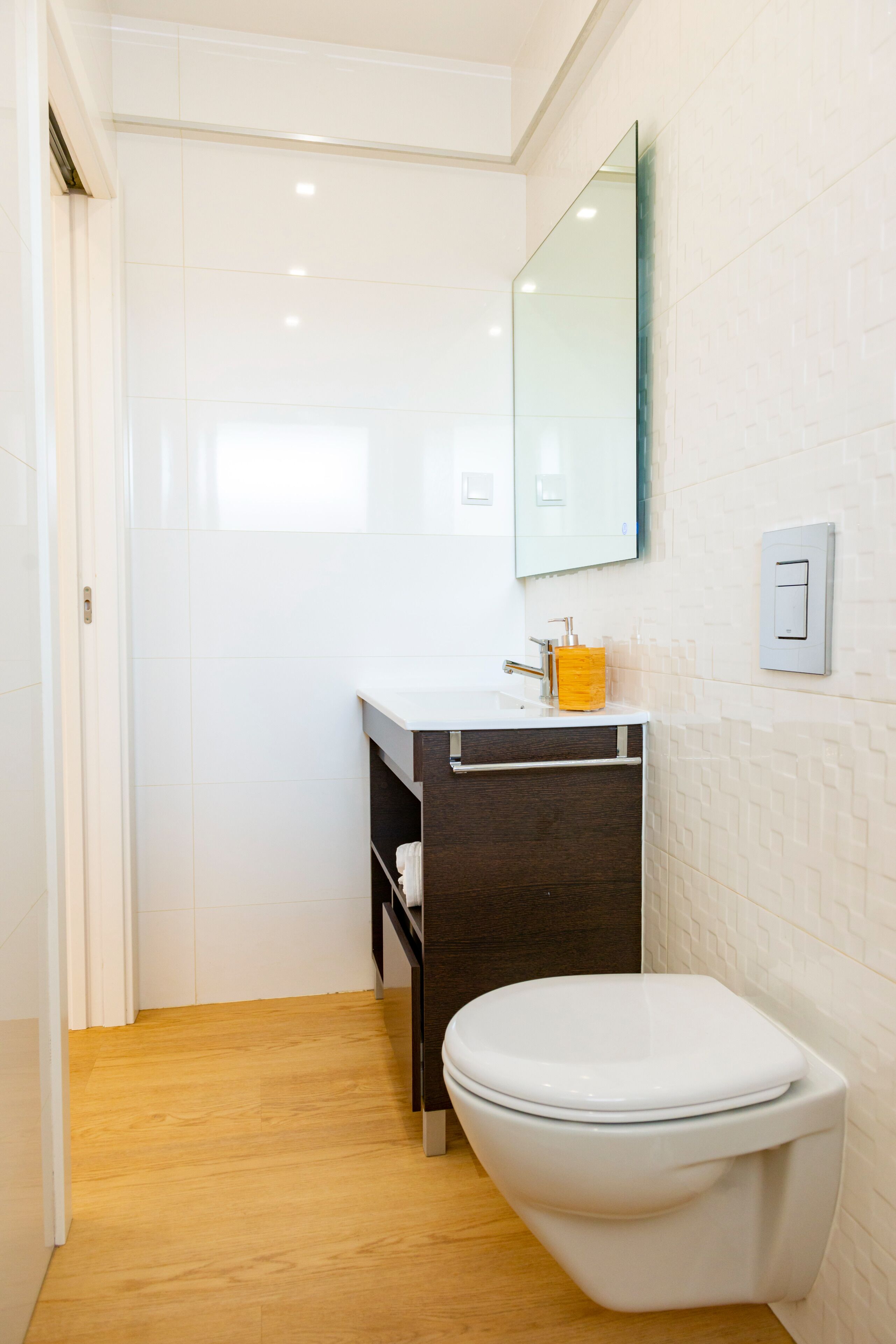 A modern bathroom with a white toilet, a wooden floor, a white sink with a dark cabinet, and a mirror on the wall.