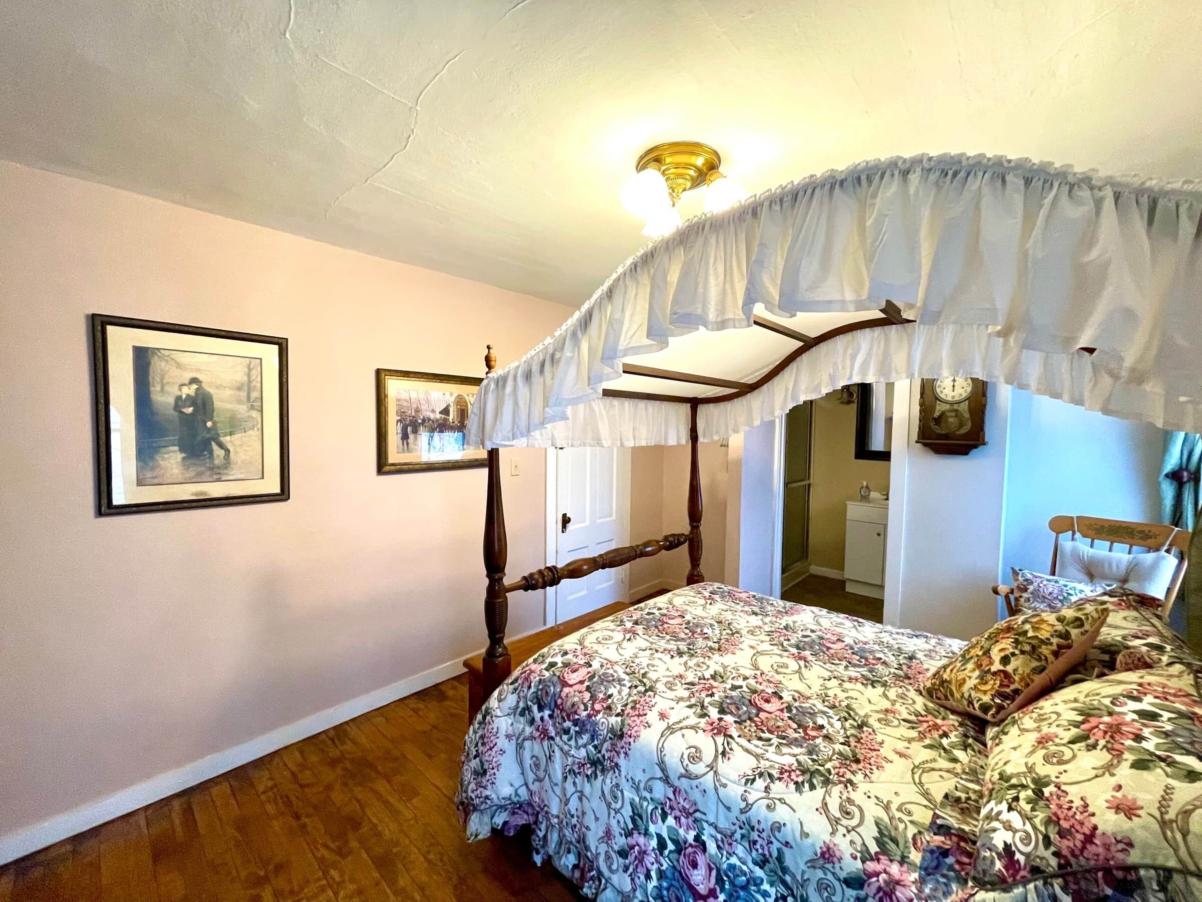 Un dormitorio con cama con dosel, ropa de cama floral, dosel, cuadros en la pared y suelo de madera.