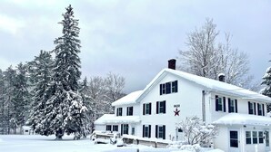 A large white house with black shutters, surrounded by snow-covered trees and a fence.
