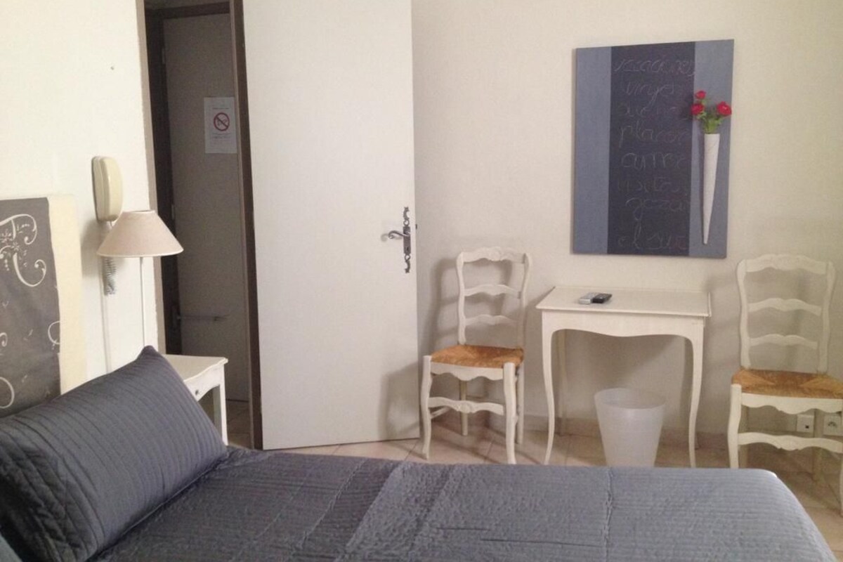 A bedroom with a bed, a desk, two chairs, a door, and a wall-mounted artwork.