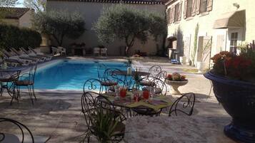 Un área junto a la piscina con sillones reclinables, una mesa de comedor para dos personas y una jardinera grande con flores.