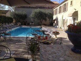 Un área junto a la piscina con sillones reclinables, una mesa de comedor para dos personas y una jardinera grande con flores.