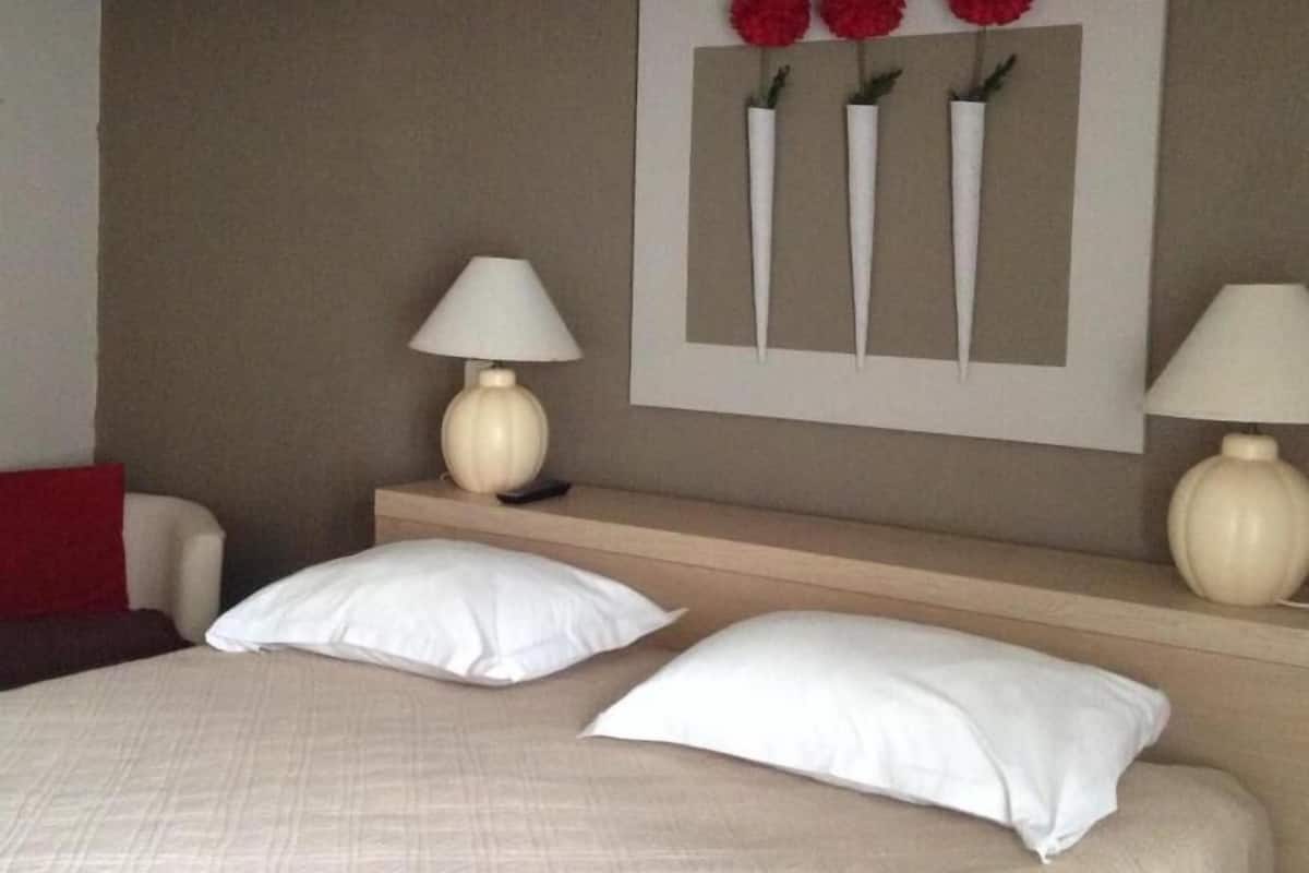 Une chambre d’hôtel avec un lit, deux tables de chevet, une lampe sur chaque table, un oreiller rouge et une décoration murale composée de trois fleurs rouges dans des vases blancs.