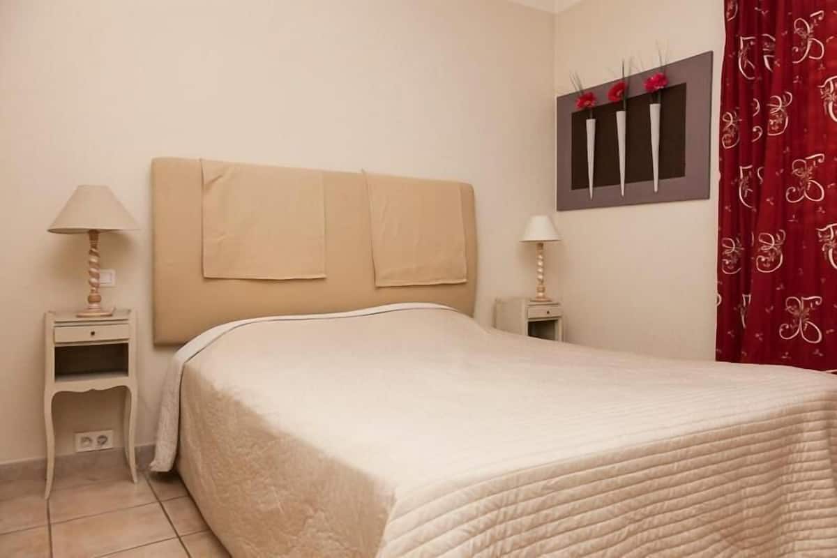 Un lit bien fait, avec une tête de lit beige, deux tables de chevet avec des lampes et une décoration murale représentant des fleurs rouges.
