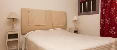 Una cama bien hecha con un cabecero color beige, dos mesitas de noche con lámparas y un adorno en la pared con flores rojas.