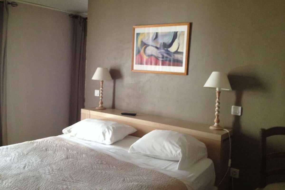 Une chambre d’hôtel avec un lit, deux lampes de chevet, un tableau abstrait encadré et une fenêtre avec des rideaux.