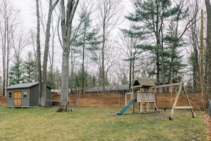 Children can play in the backyard playhouse with a slide, surrounded by a grassy lawn and a wooden fence.