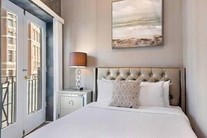 A hotel room with a large bed, a bedside table with a lamp, and a view of the city through a door.