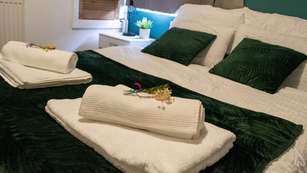 A neatly made bed with white linens and green pillows, a small bedside table with a lamp and a potted plant, and a window with blinds. - 2 bed house Sedley Street Liverpool (Liverpool)