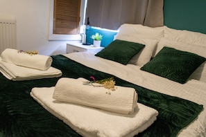 A neatly made bed with white linens and green pillows, a small bedside table with a lamp and a potted plant, and a window with blinds.