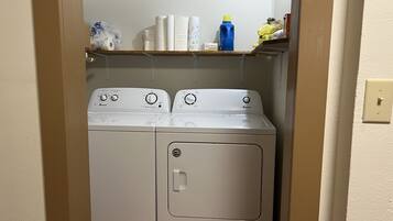 Una unidad compacta de lavadora y secadora con panel de control, ubicada en un lavadero pequeño con un estante superior que contiene diversos productos de limpieza.