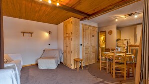 Ein Zimmer mit einem Bett, einem kleinen Hocker, Holzschränken und einem Essbereich mit Tisch und Stühlen.