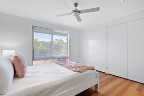 A bedroom with a bed, a ceiling fan, a window with a view, and white wardrobes.