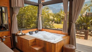 Uma banheira de madeira com escadas, um banquinho pequeno e vista para um jardim exuberante e montanhas através de grandes janelas.