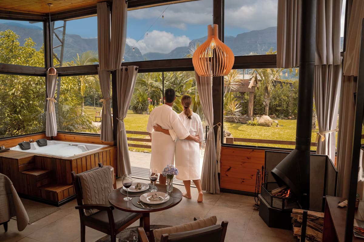 A couple standing by a large window overlooking a lush garden and mountains.