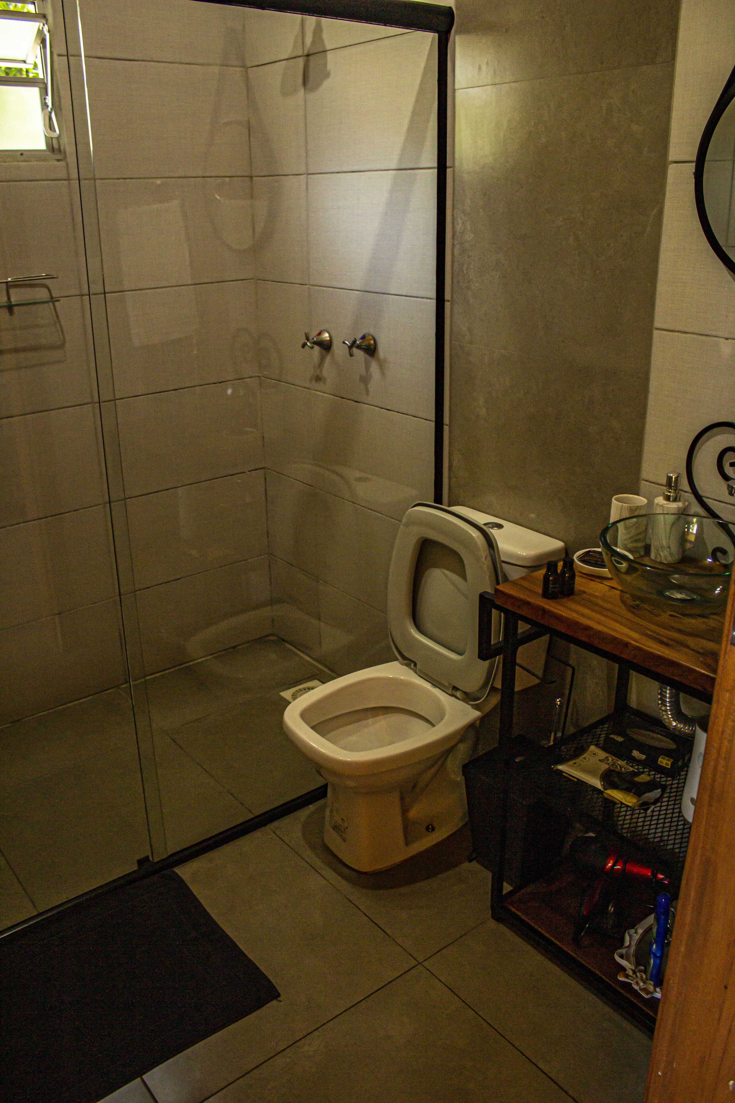 Une salle de bain comprenant des toilettes, une douche à parois de verre et une étagère en bois où sont rangés les articles de toilette.