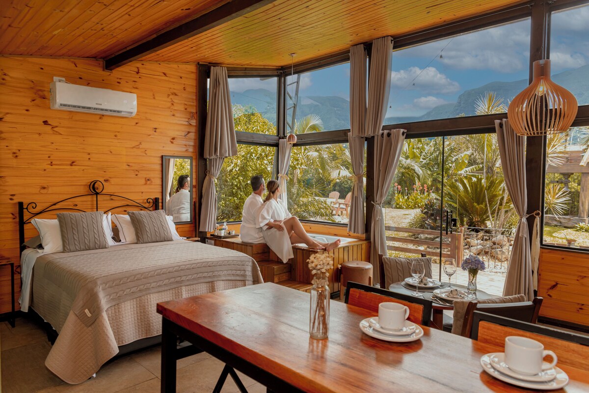 A wooden room with a bed, a dining table with cups and a vase, and a view of mountains and greenery.