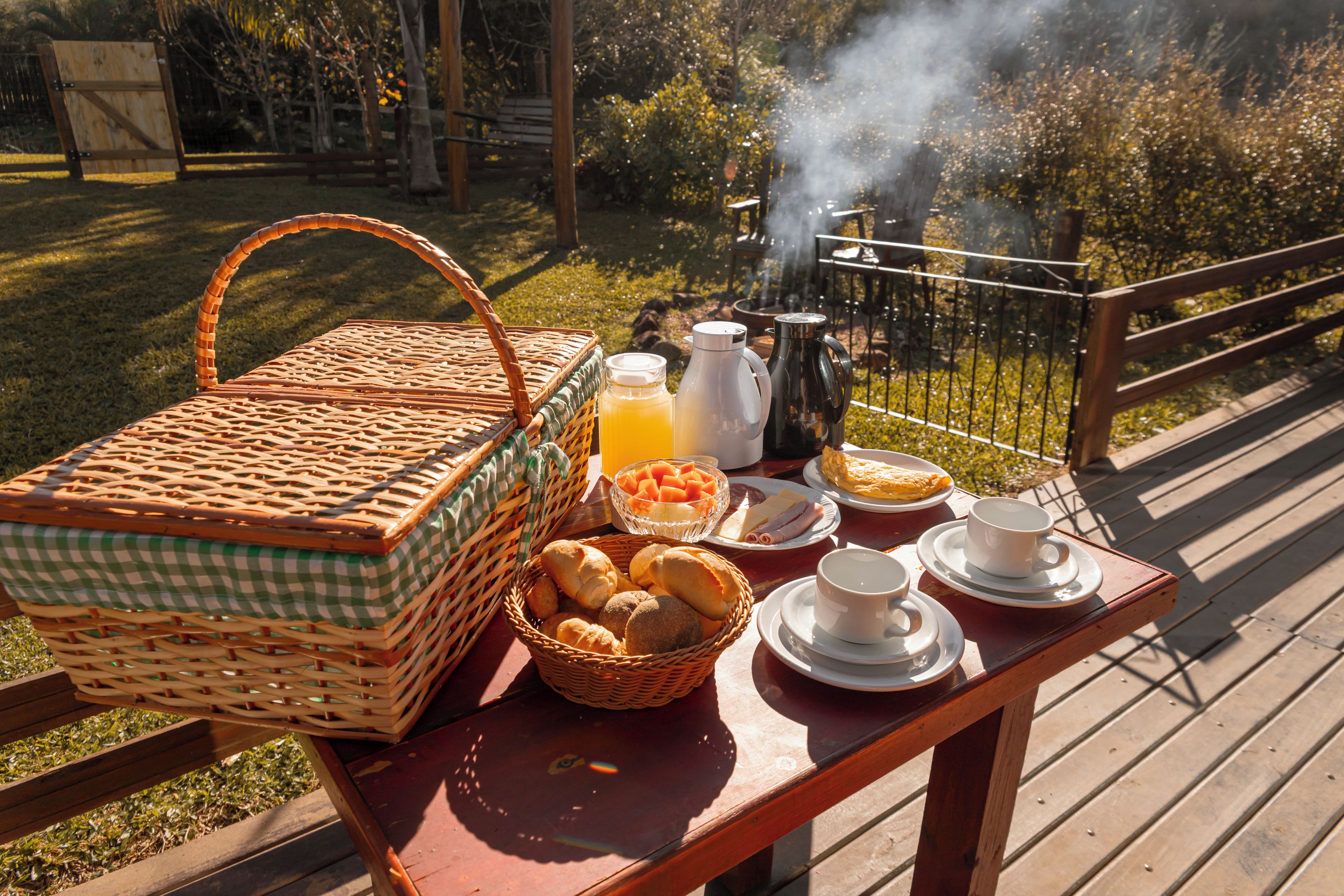 Un montaje para picnic con una cesta de mimbre, un cuenco de fruta, una botella de jugo y una cafetera sobre una mesa de madera.