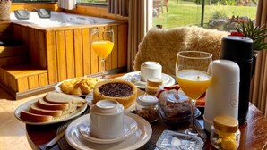 Mesa de madeira com comida, suco de laranja e máquina de café, em um cenário com vista para montanhas.