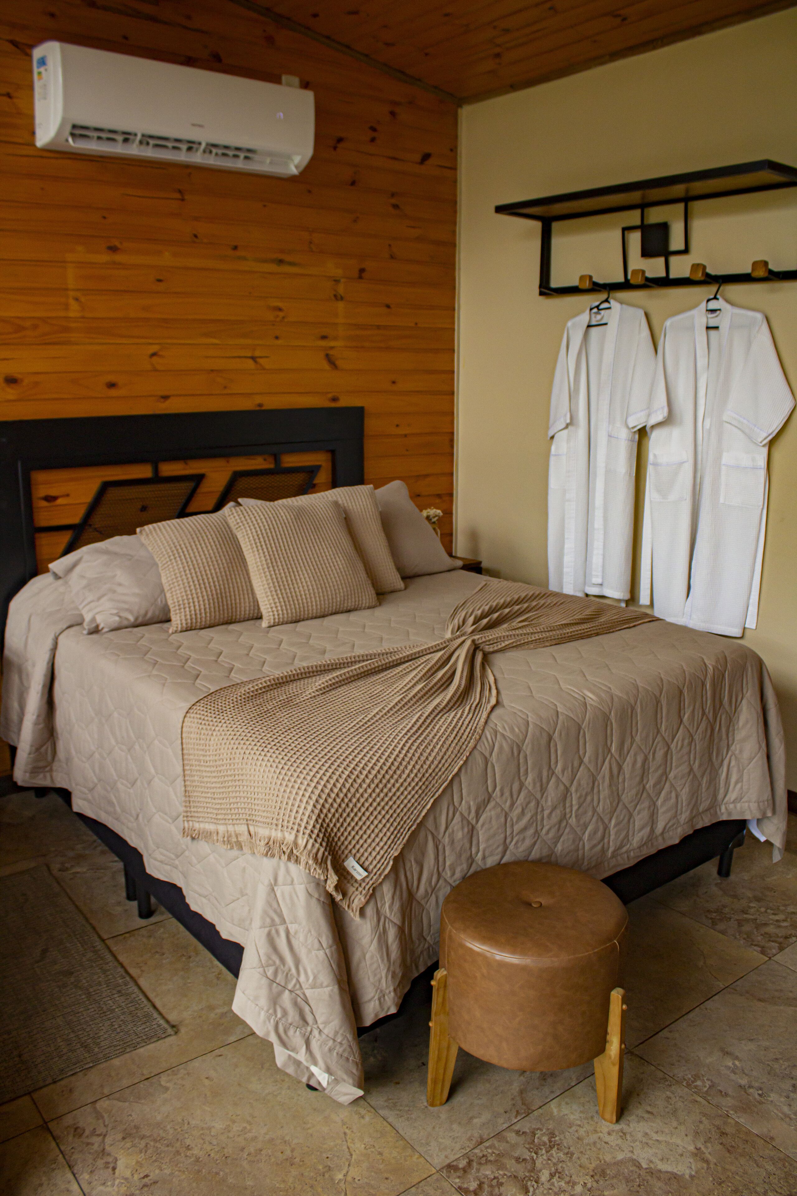 A bedroom with a bed, a wooden stool, and a wall-mounted rack with clothes.