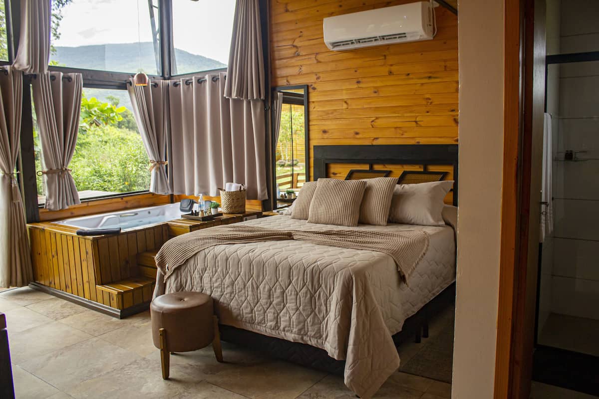 A bedroom with a wooden headboard, a large window with curtains, a built-in bathtub, and a bedside table with a lamp.