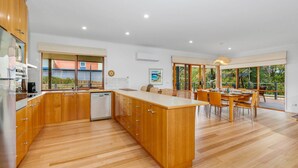 Una cocina moderna con armarios de madera, lavavajillas y una isla grande. Hay una zona de comedor con una mesa y sillas, y ventanas amplias con vistas al exterior.