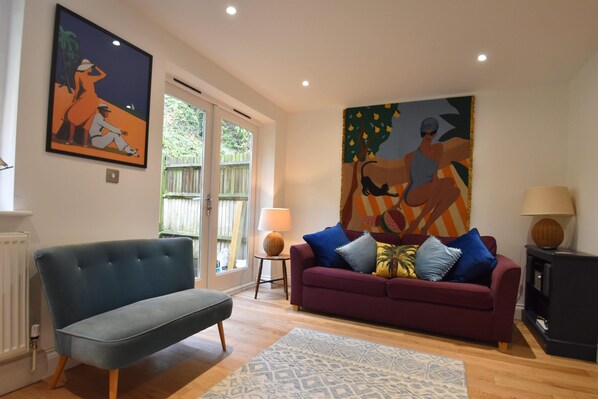 A living room with a blue sofa, a purple sofa with multiple cushions, a patterned rug, a painting on the wall, and a lamp on a side table. - Southbank family-friendly holiday cottage in Rye, close to the centre (Rye)