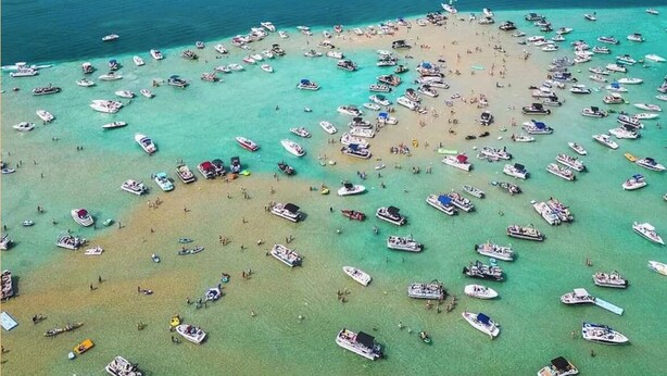 Vista aérea de una playa con numerosas embarcaciones ancladas en aguas poco profundas, rodeada de abundante vegetación y un cielo despejado.
