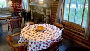 Un interior de cabaña acogedor con una mesa de comedor dispuesta para cuatro personas, una chimenea de piedra, un televisor montado en la pared y una zona de estar cómoda con un sofá y sillones.