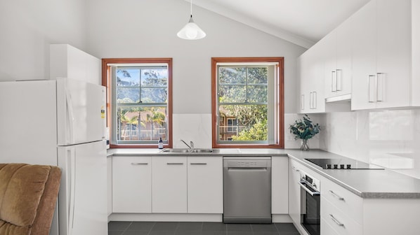 Eine moderne Küche mit weißen Schränken, einer Edelstahl-Spüle und einem Geschirrspüler. Zwei Fenster mit Holzrahmen sowie eine Deckenleuchte sind vorhanden.
