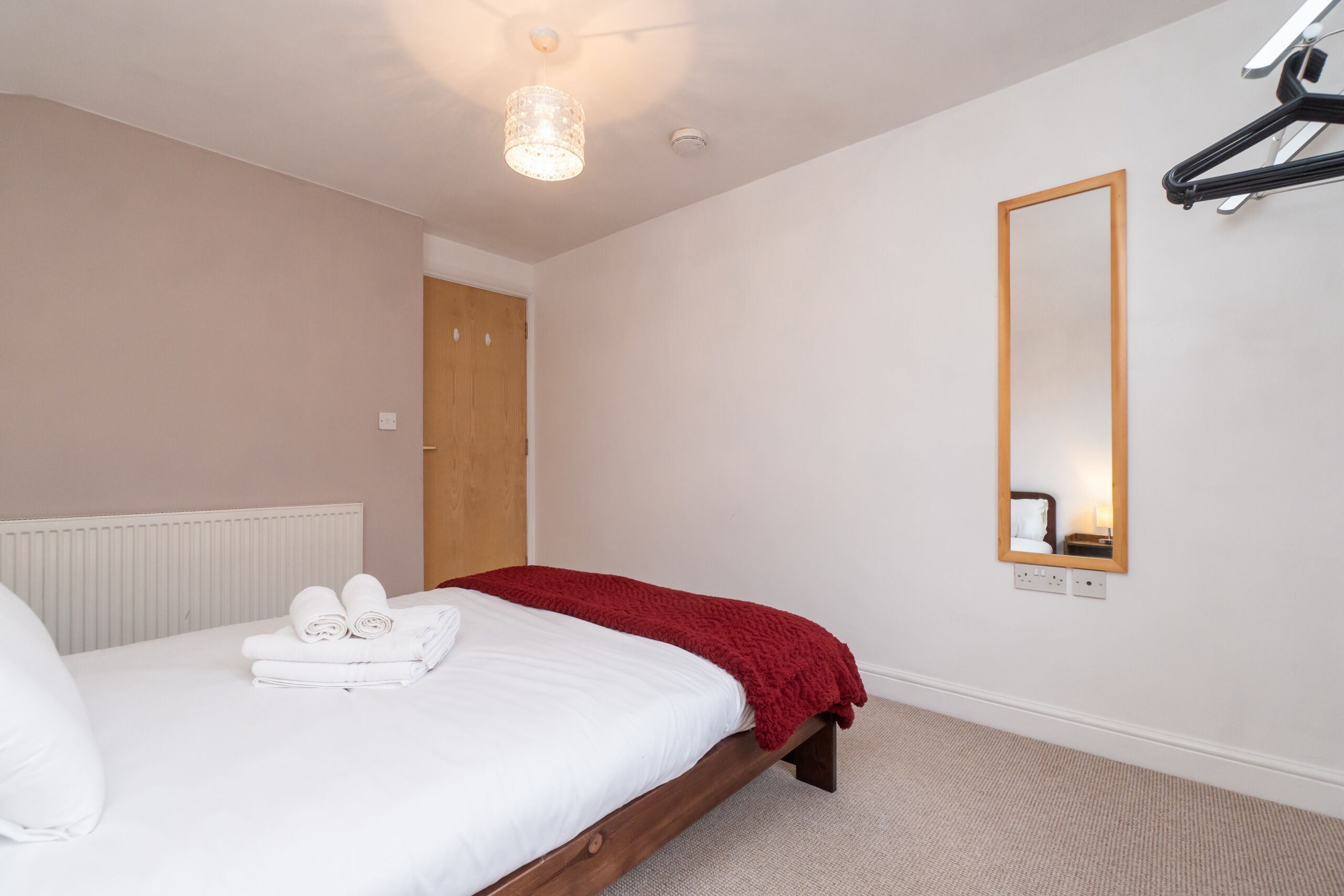 A neatly made bed with white linens and a red blanket, a wooden headboard, a hanging light fixture, a mirror on the wall, and a door in the background.