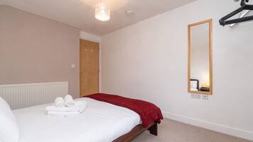 A neatly made bed with white linens and a red blanket, a wooden headboard, a hanging light fixture, a mirror on the wall, and a door in the background.
