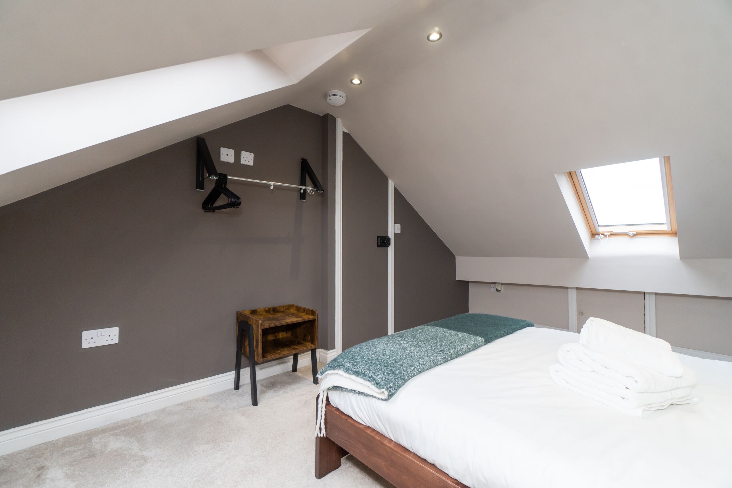 Un dormitorio con techo inclinado, tragaluz, cama con sábanas blancas impecables, mesita de noche de madera y perchero en la pared.