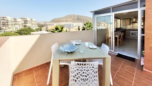 Un balcón con mesa y sillas, una puerta corrediza de vidrio que da a una cocina, y vistas de un paisaje urbano con edificios y una montaña al fondo.