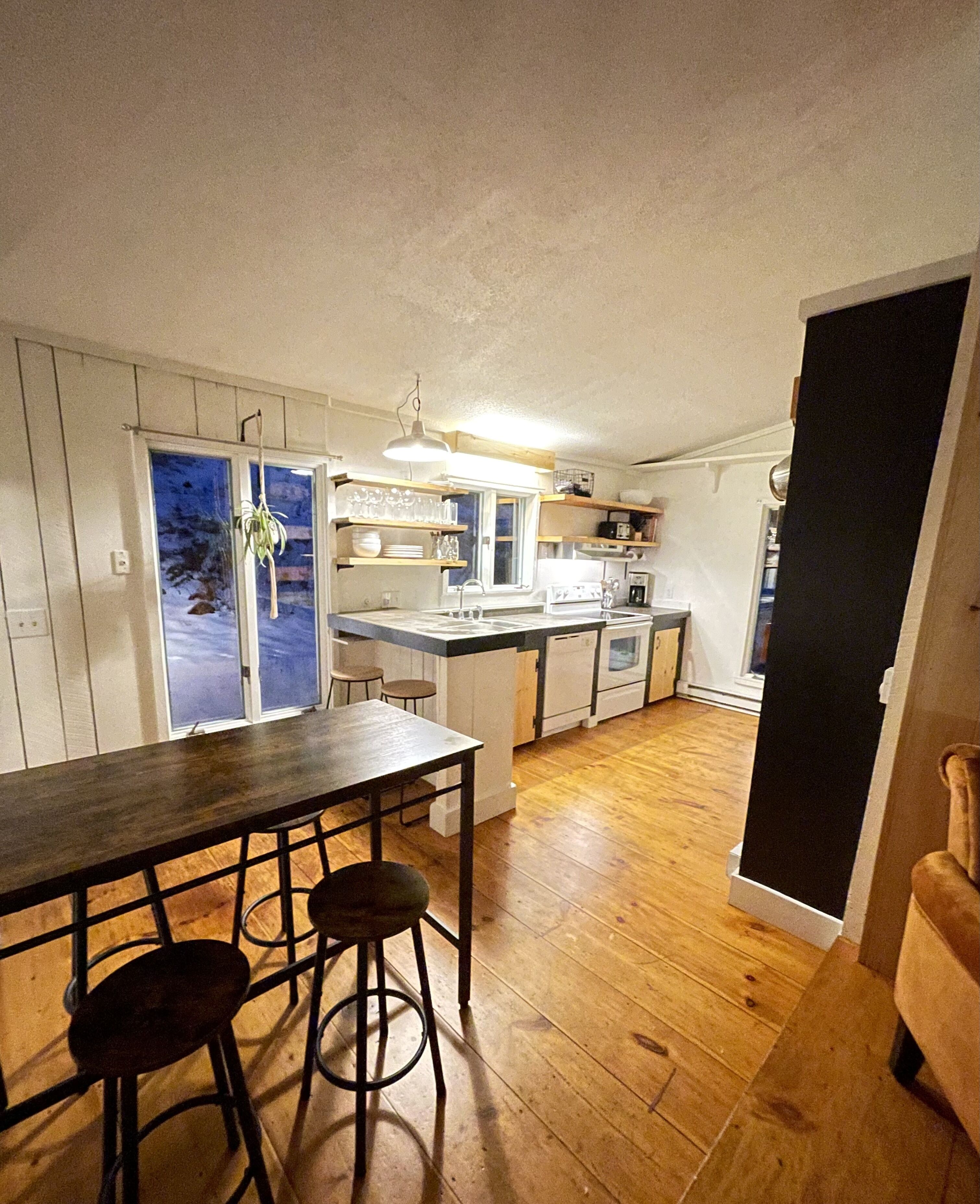 A compact kitchen with wooden flooring, a dining table with stools, and a view through a window.