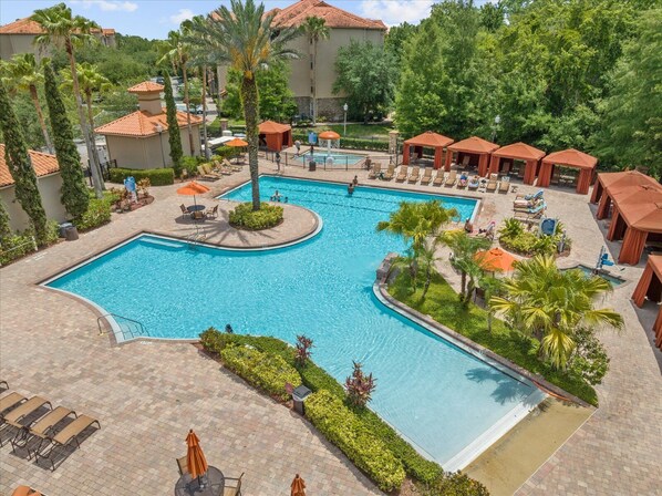 A large outdoor swimming pool with multiple lounge areas, palm trees, and a building with a tiled roof in the background. - Near Disney w/ pool-3BR/2BA-Spacious & Cozy Condo (Davenport)