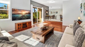 Una sala moderna con un televisor de pantalla plana grande, una mesa de centro de madera y un sofá. Hay una puerta corrediza de vidrio que da a un balcón con vistas a zonas verdes.