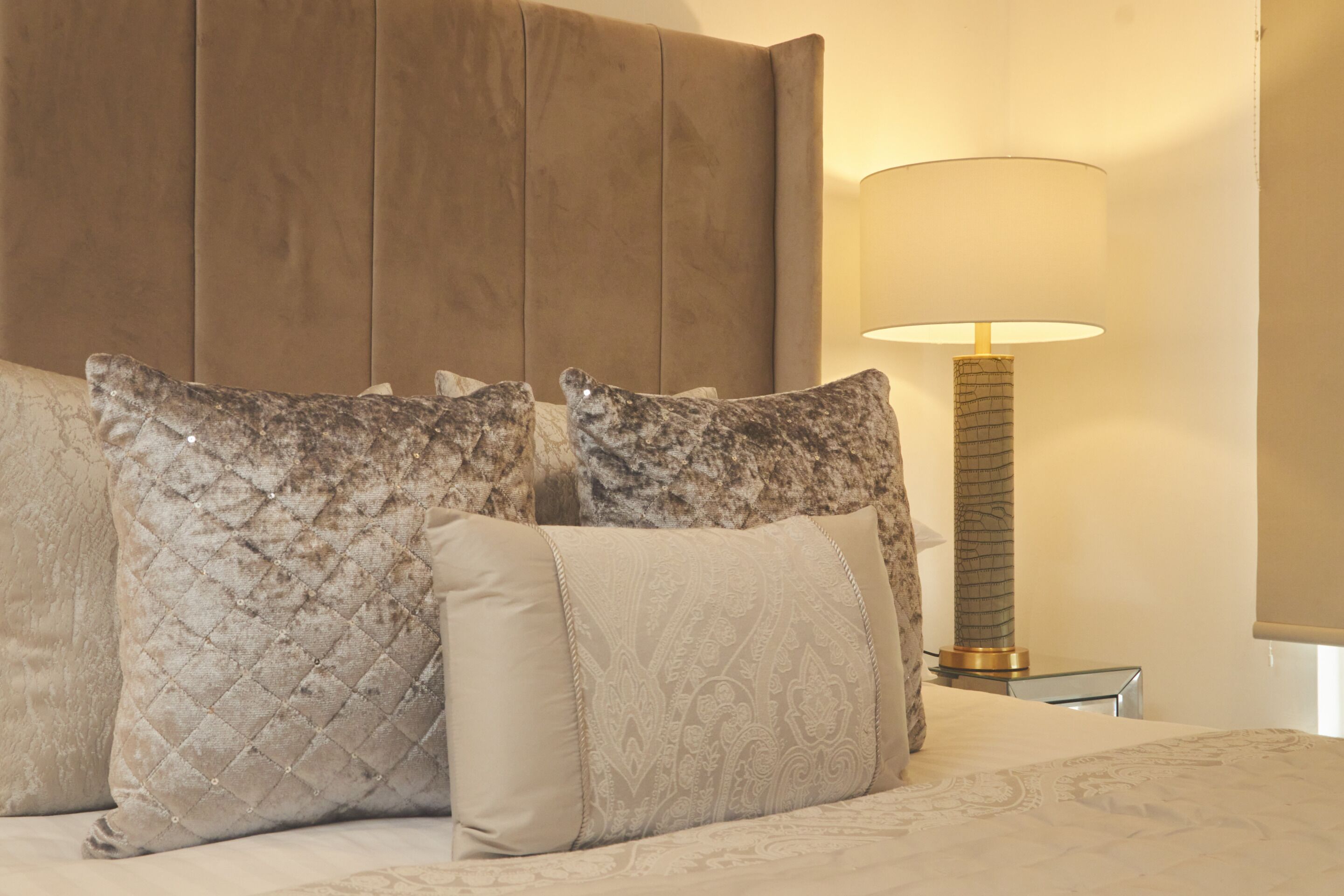 A neatly made bed with patterned pillows and a headboard, a bedside lamp, and a glass table beside the bed.