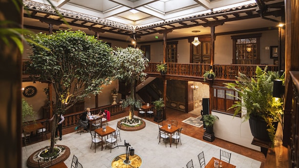An indoor atrium with a high ceiling, wooden beams, and a central tree. Tables and chairs are arranged around the space, with potted plants and a fireplace.