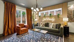 Ein Wohnzimmer mit Sofa, Sessel, Couchtisch und einem gemusterten Teppich. Ein Fenster mit Vorhängen, eine Neonreklame und eine Wandpflanze sind ebenfalls vorhanden.