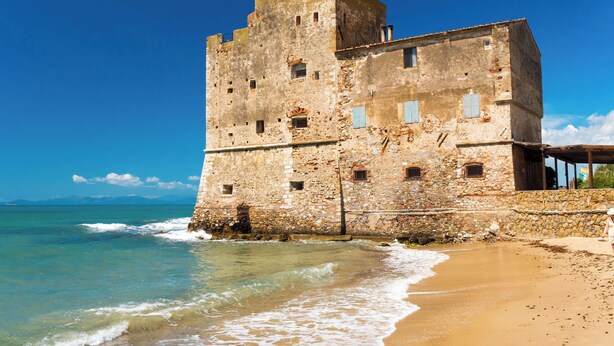 Um edifício histórico de pedra, com várias janelas e uma torre, localizado à beira de uma praia com águas azul-claras.