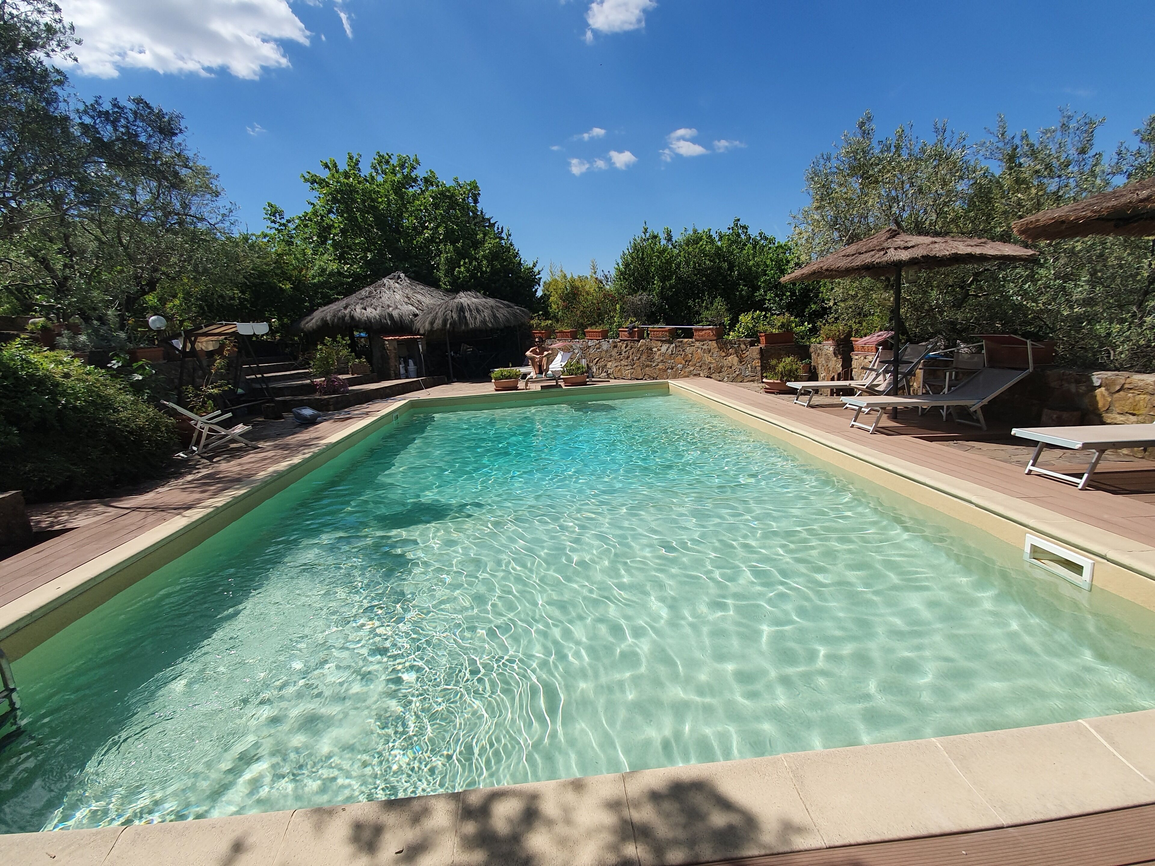 Piscina rectangular amb aigua cristal·lina, envoltada de gandules i ombrel·les, situada en un jardí paisatjístic amb arbres i una paret de pedra.