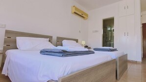 A hotel room with two beds, a wooden headboard, white bedding, a wall-mounted air conditioner, a nightstand with a lamp, and a wardrobe. - STAY Nissi Heaven (Ayia Napa)