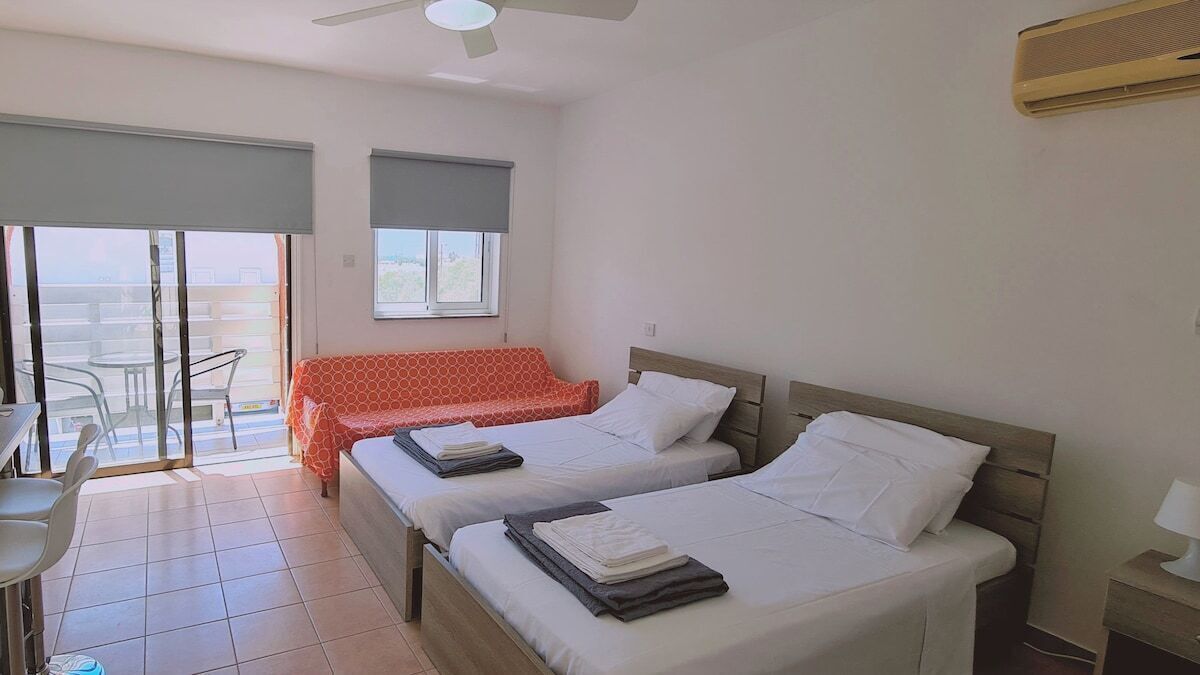 A hotel room with two beds, a balcony with a table and chairs, and a ceiling fan.