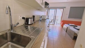A modern kitchen with a stainless steel sink, a kettle, a toaster, and a microwave. There is a balcony with a glass door and a bed with an orange headboard. - STAY Nissi Heaven (Ayia Napa)