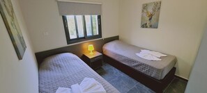 Ein Zimmer mit zwei Betten, einem Nachttisch mit Lampe, einem Fenster mit Jalousien und einem Bild an der Wand.